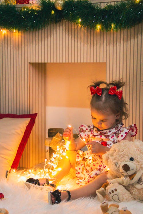 A Young Girl in Printed Dress Sitting Near Her Plush Toy while Holding String Lights