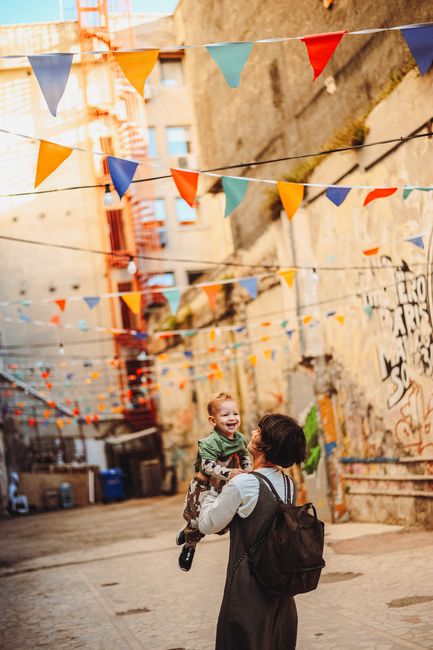 A mother lifts her smiling child in a vibrant alley with colorful flags and street art.