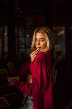 Stylish blonde woman in red jacket poses alluringly in a dimly lit room.