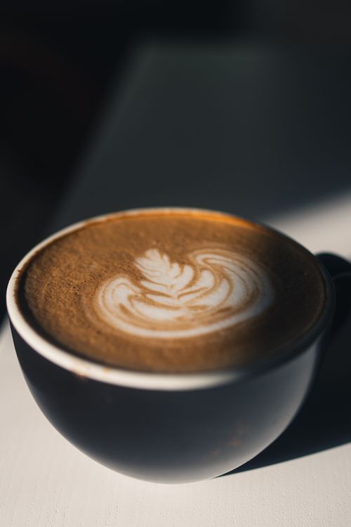 Free A close-up of a latte with intricate frothy art, captured in warm sunlight casting deep shadows. Stock Photo