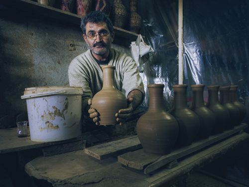 Free A craftsman in a workshop carefully shapes a clay vase by hand, surrounded by finished pottery. Stock Photo