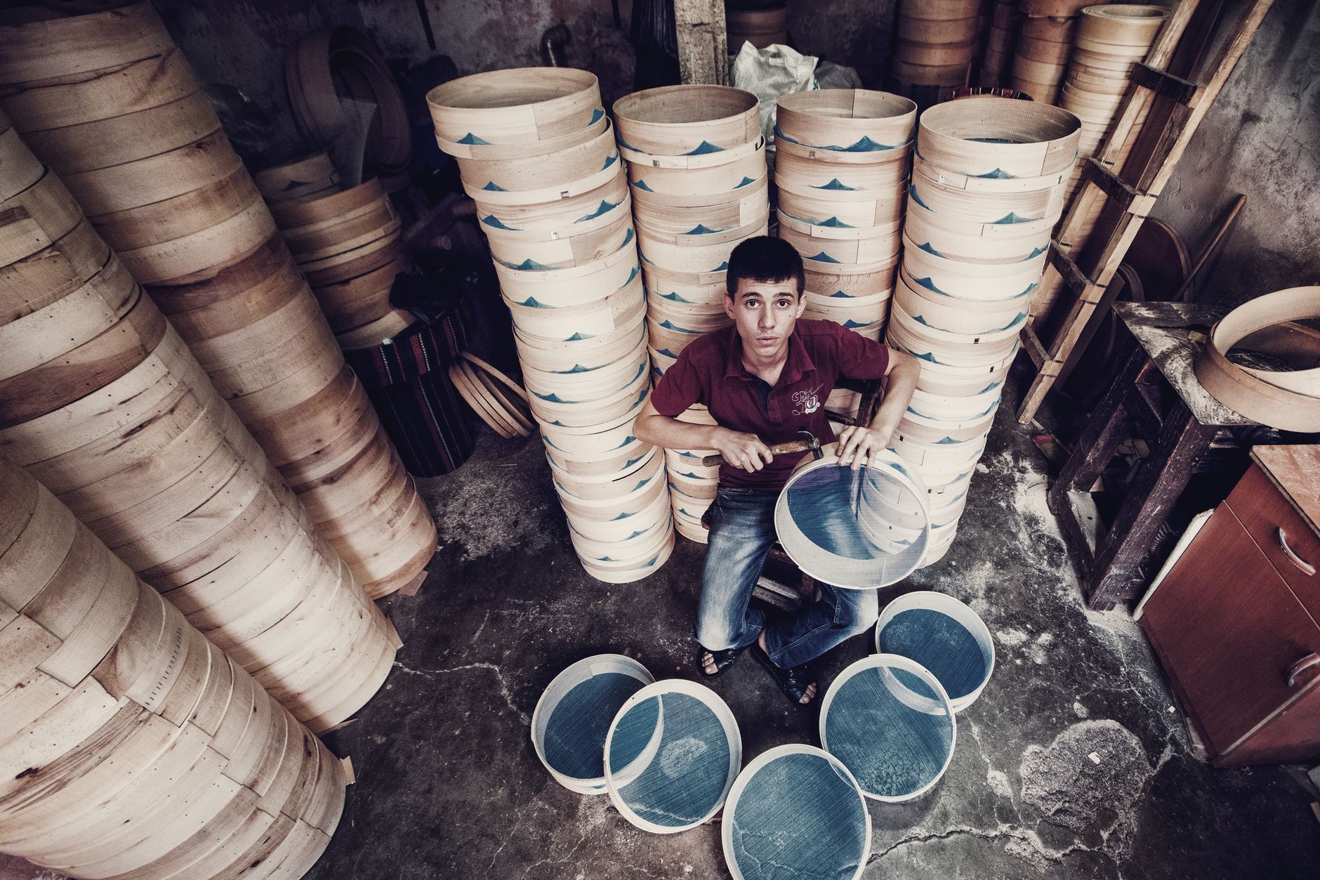 Portrait of a young artisan crafting sieves in a traditional workshop surrounded by tall stacks of wooden frames.