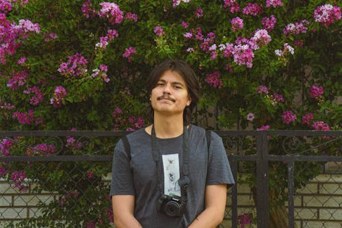 Man with Mustache Wearing a Camera on a Strap and Standing under a Shrub with Purple Flowers