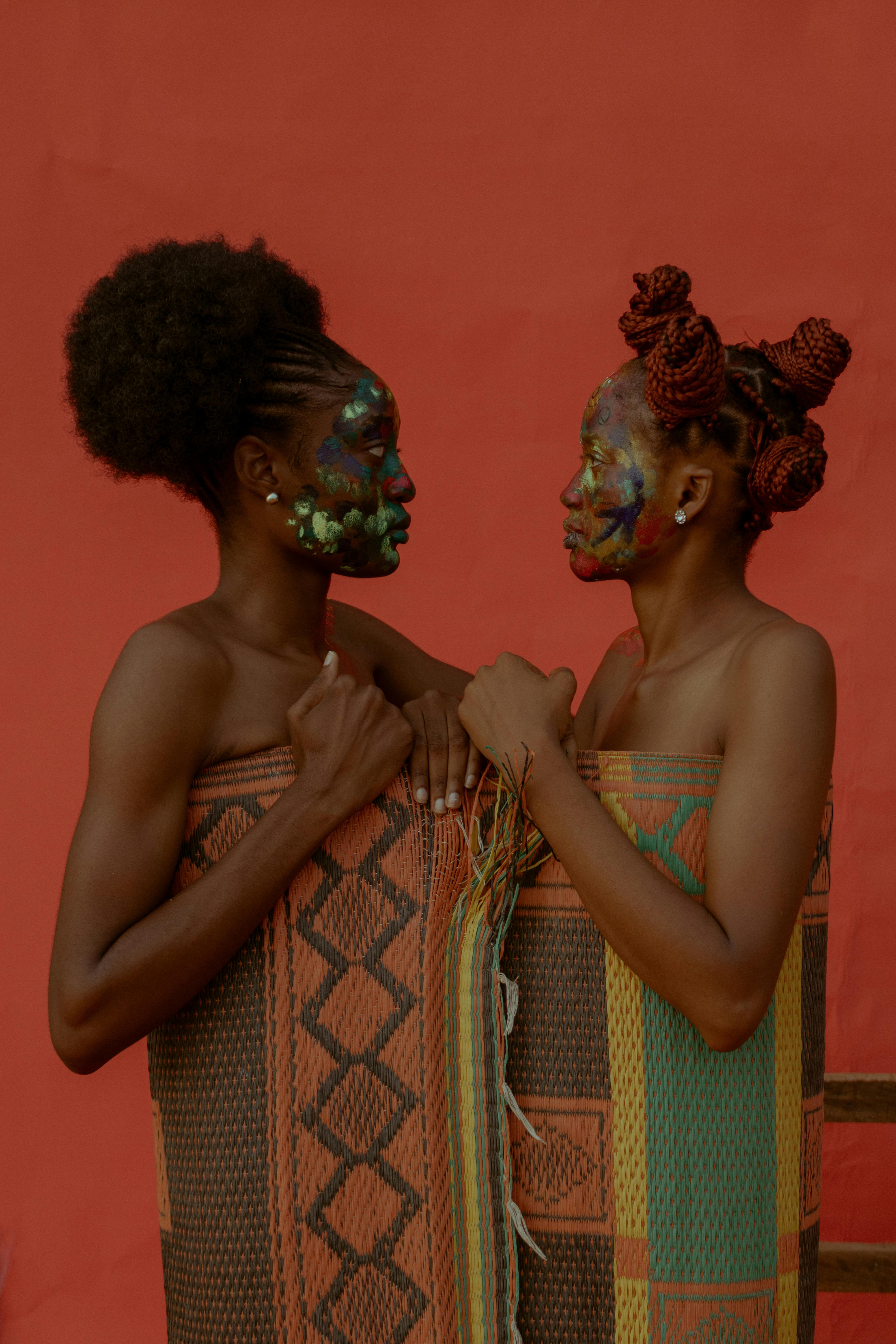 Two women with colorful face paint wrapped in traditional textiles against a red background.