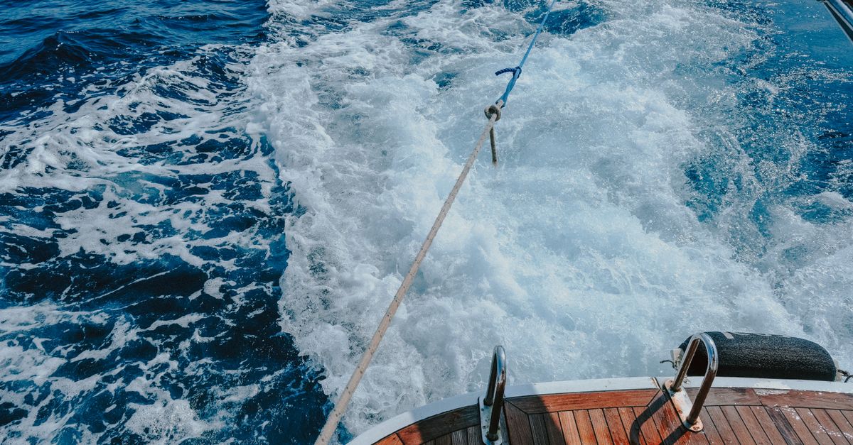 A boat's wooden deck with a rope trailing in the sea leaving a water trail behind.