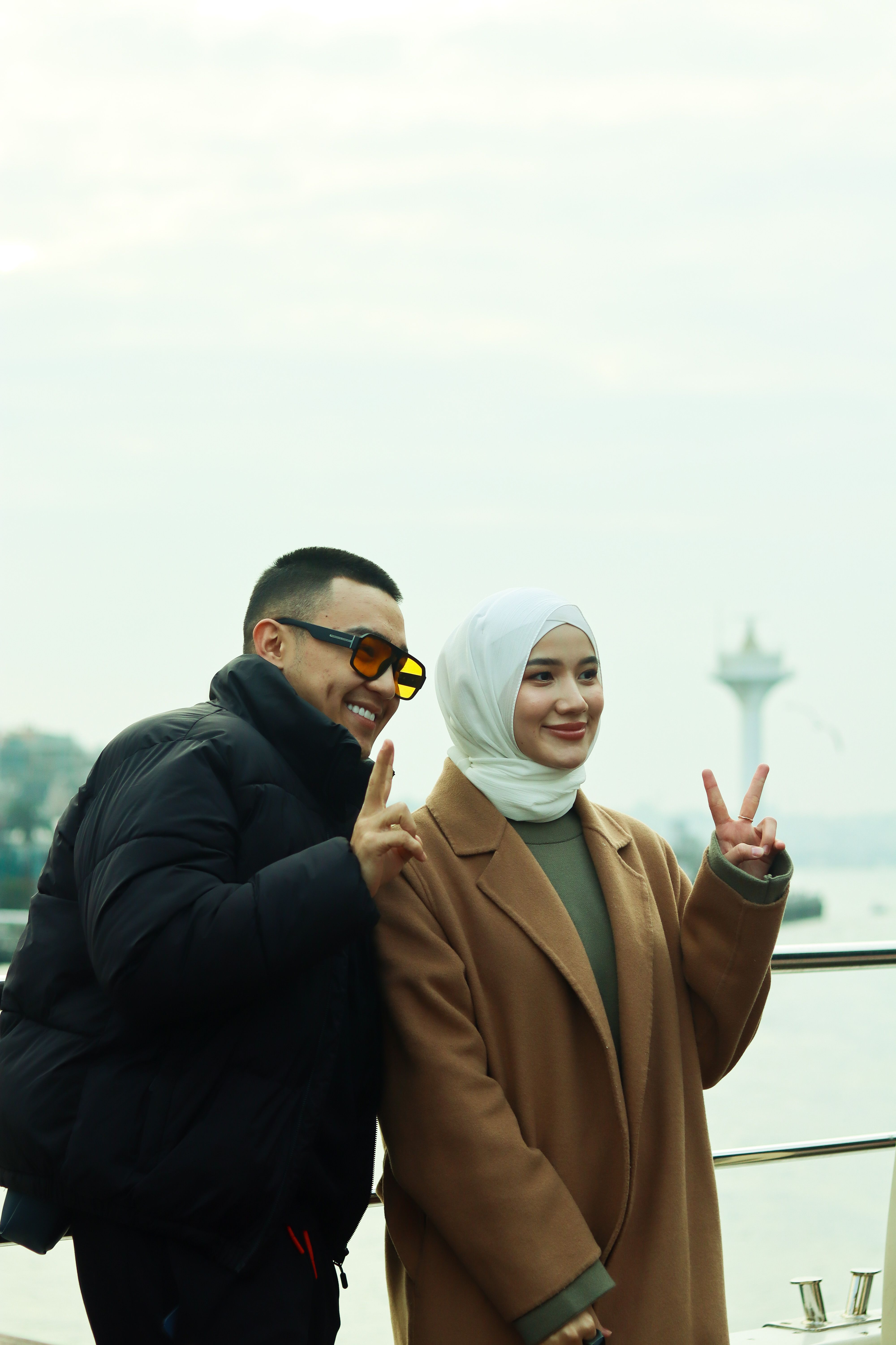 A joyful couple poses with peace signs on a waterfront, capturing a vibrant moment.