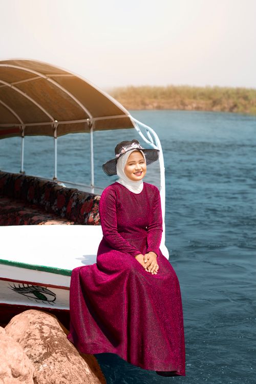 Free A smiling young woman in a floral hat enjoys a sunny day by the tranquil water, sitting on a boat deck. Stock Photo