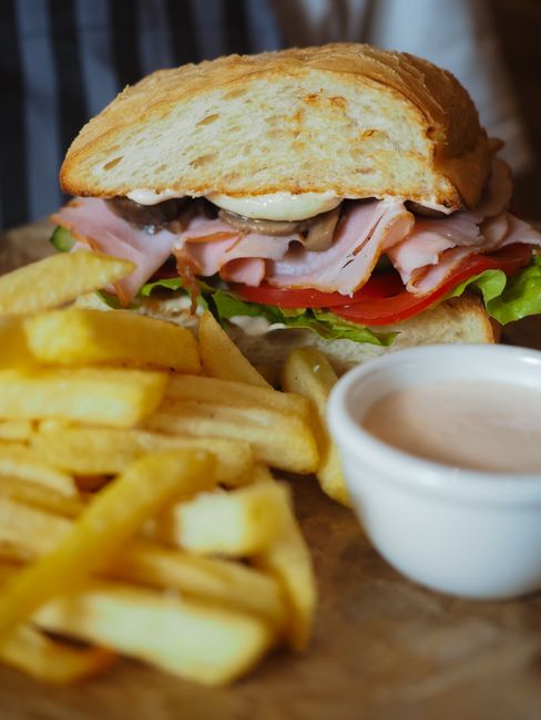 A sliced ham sandwich on a wooden board, layered with deli ham, cheese, lettuce, and tomato between two slices of bread with a pickle spear on the side.
