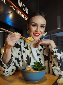 Smiling woman in printed attire holding noodles with chopsticks indoors.
