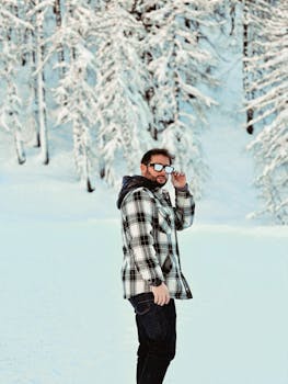 Adult man wearing sunglasses and plaid jacket standing in a snowy winter landscape.