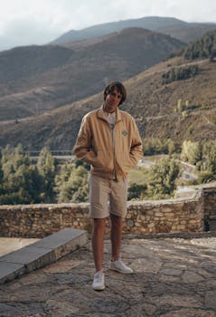 A young man in casual attire stands in a picturesque mountain setting, surrounded by hills and sunlight.