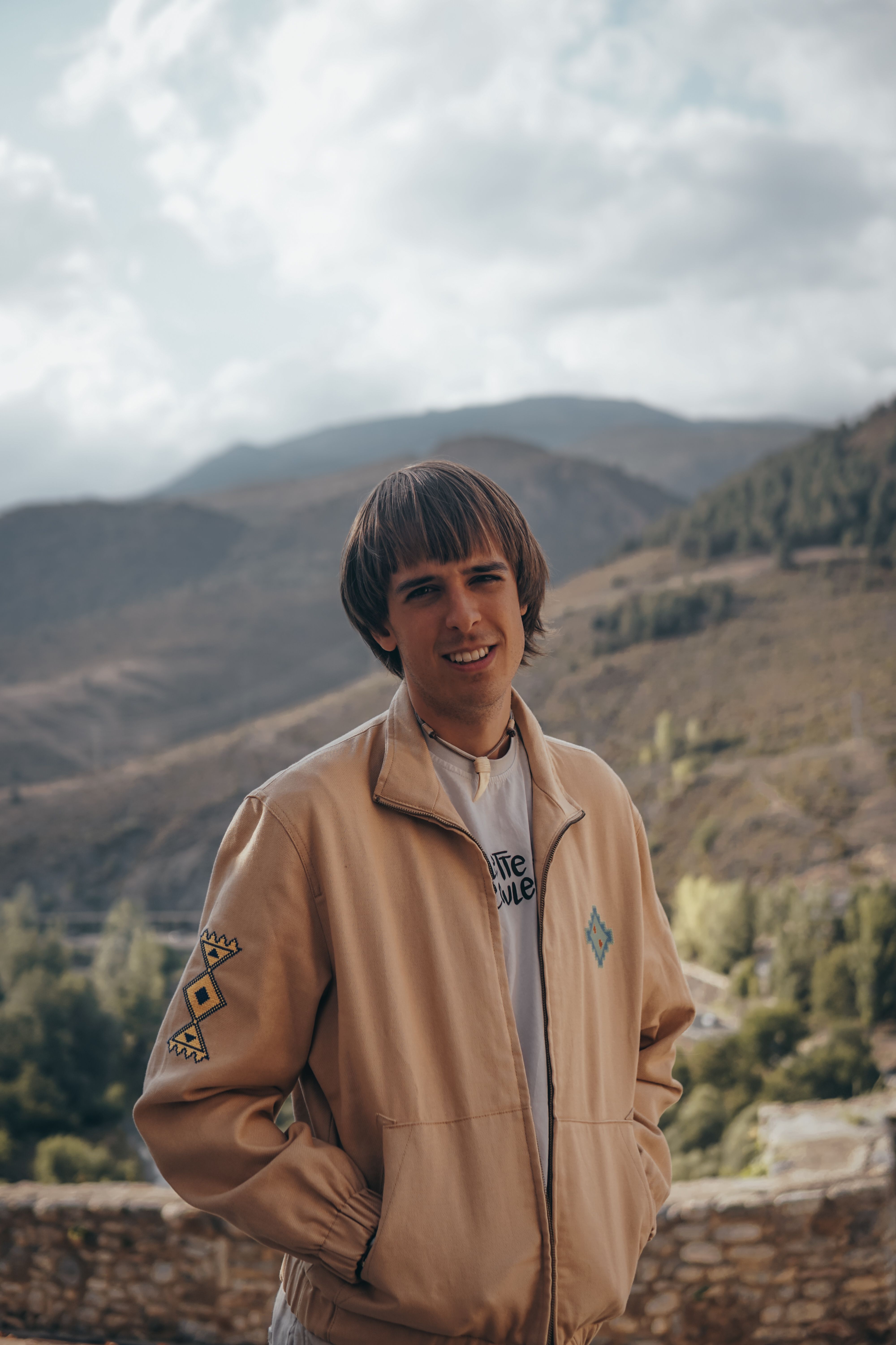 Young man posing with hands in jacket pockets against a scenic mountainous backdrop.