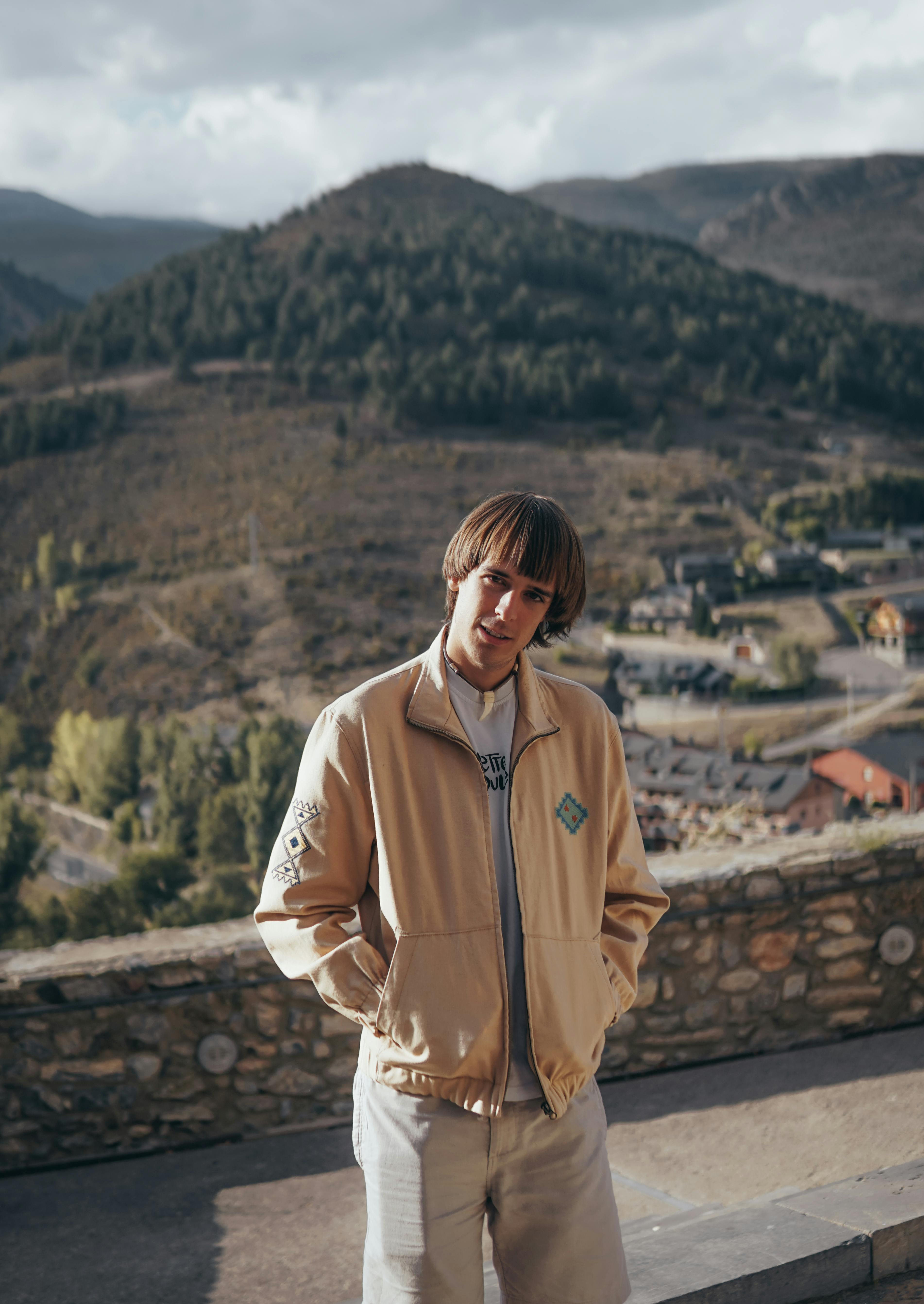 A young man in a beige jacket poses outdoors against a scenic mountain view.