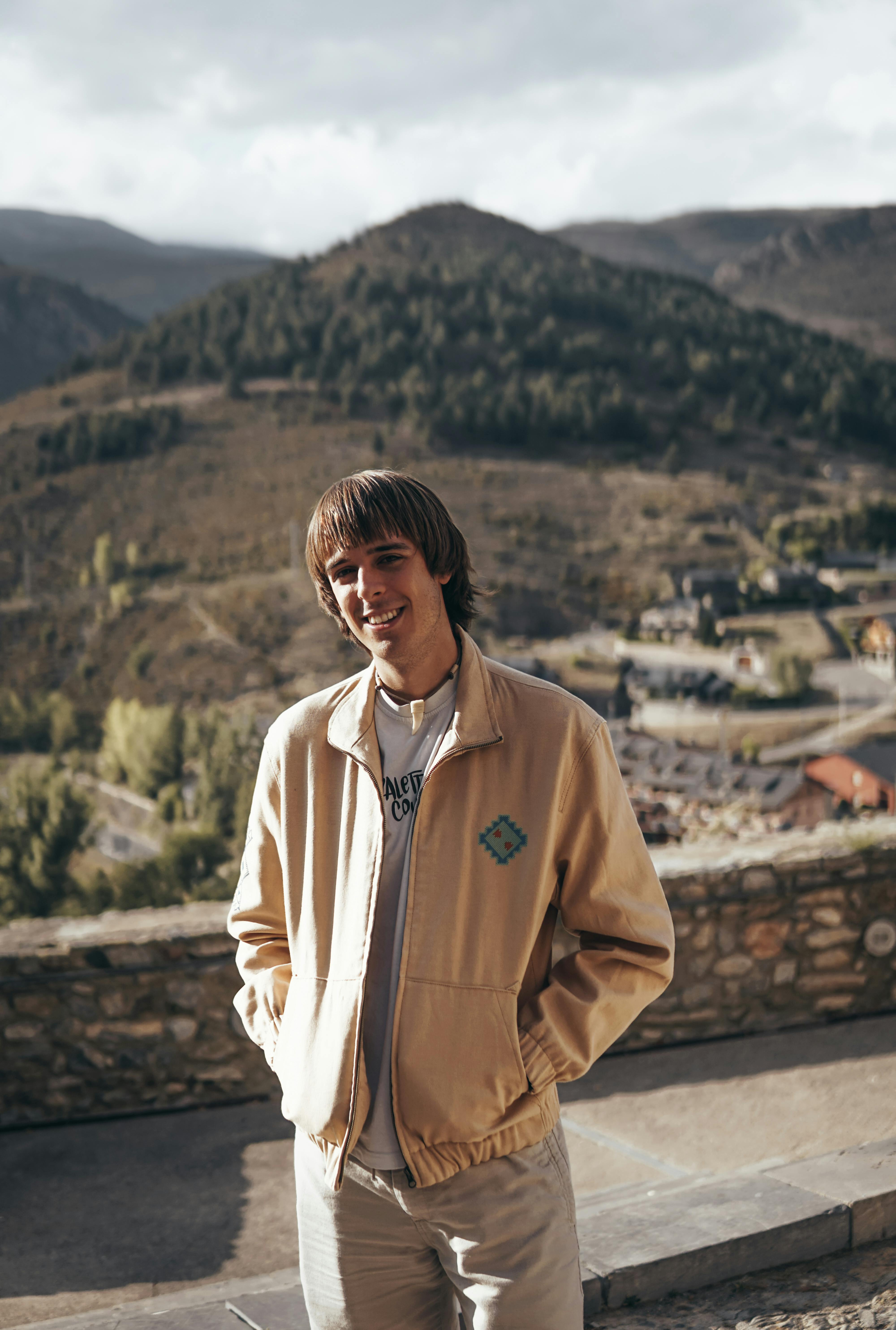 Young man smiling in a beige jacket with scenic mountain backdrop.