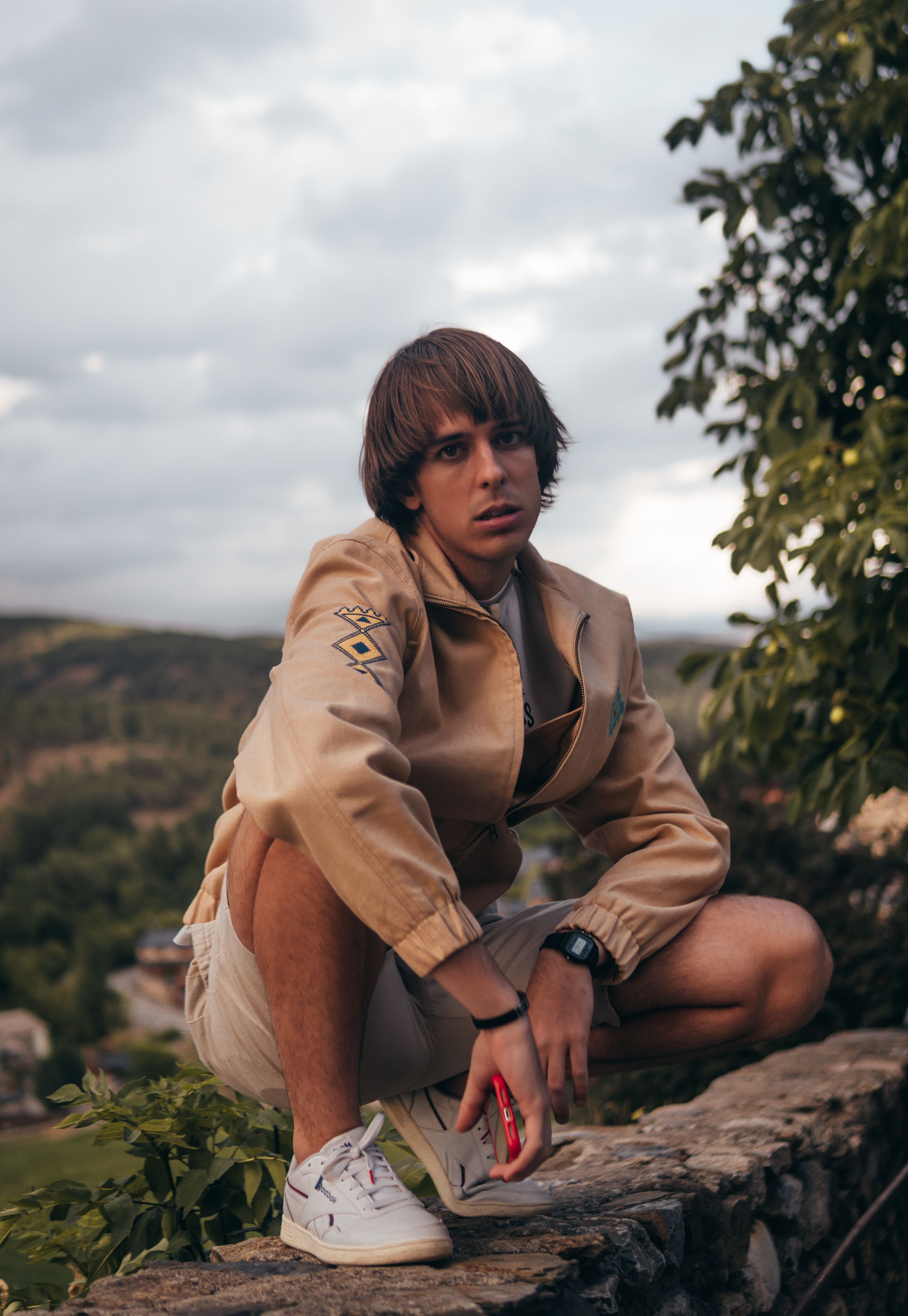 A young man crouches on a stone wall in a scenic outdoor setting, showcasing a thoughtful summer fashion pose.