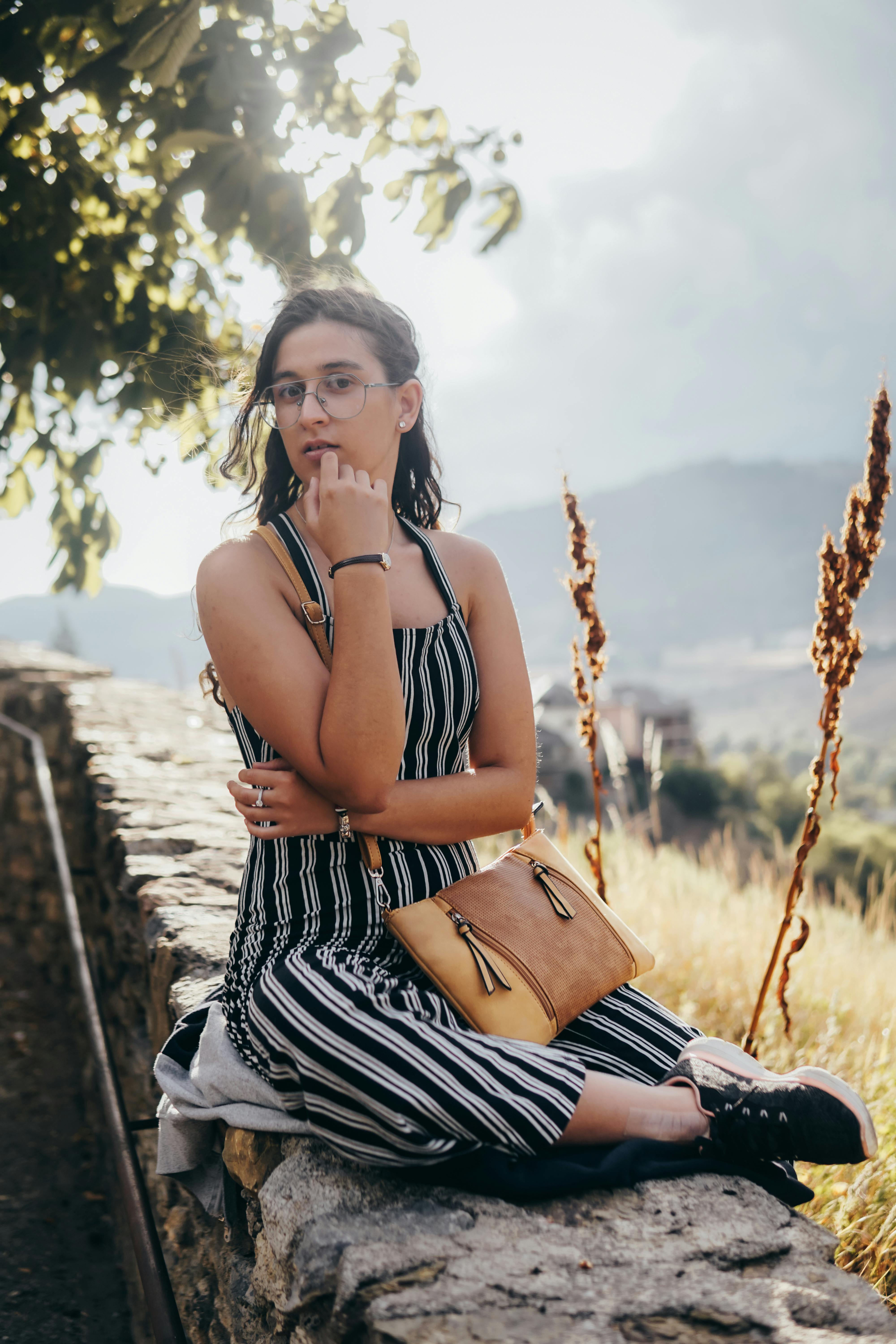 A young woman in a striped dress sits on a stone wall outdoors, enjoying the summer landscape.