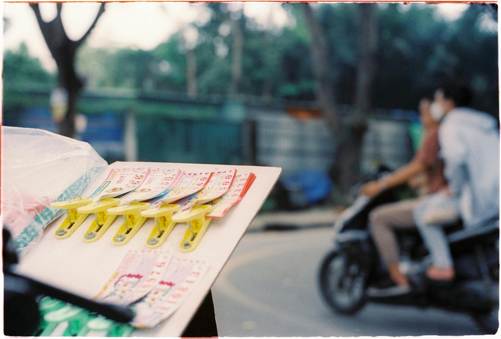 A collection of lottery tickets clipped onto a board, with people passing on a motor scooter in the blurred background.