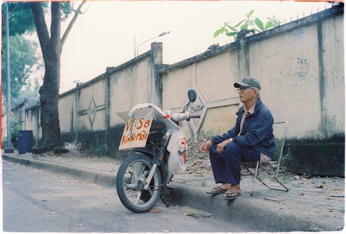Elderly Man Sitting Next to Motorbike on Street