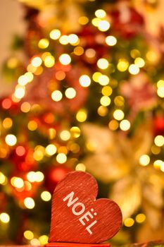 Heart-shaped ornament with 'Noël' text in front of blurred Christmas lights and tree.