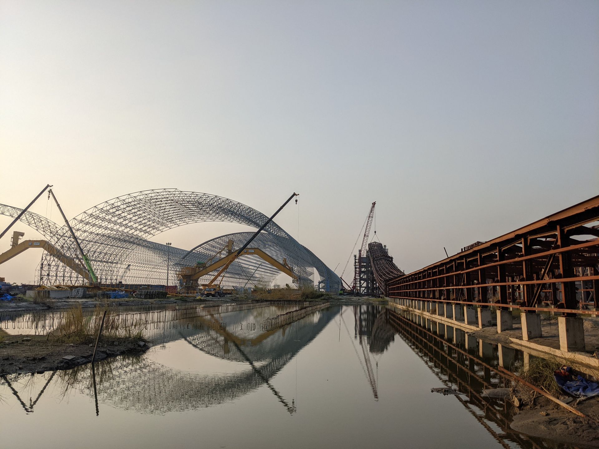 A picturesque industrial construction site with cranes at sunset, reflecting in calm water.
