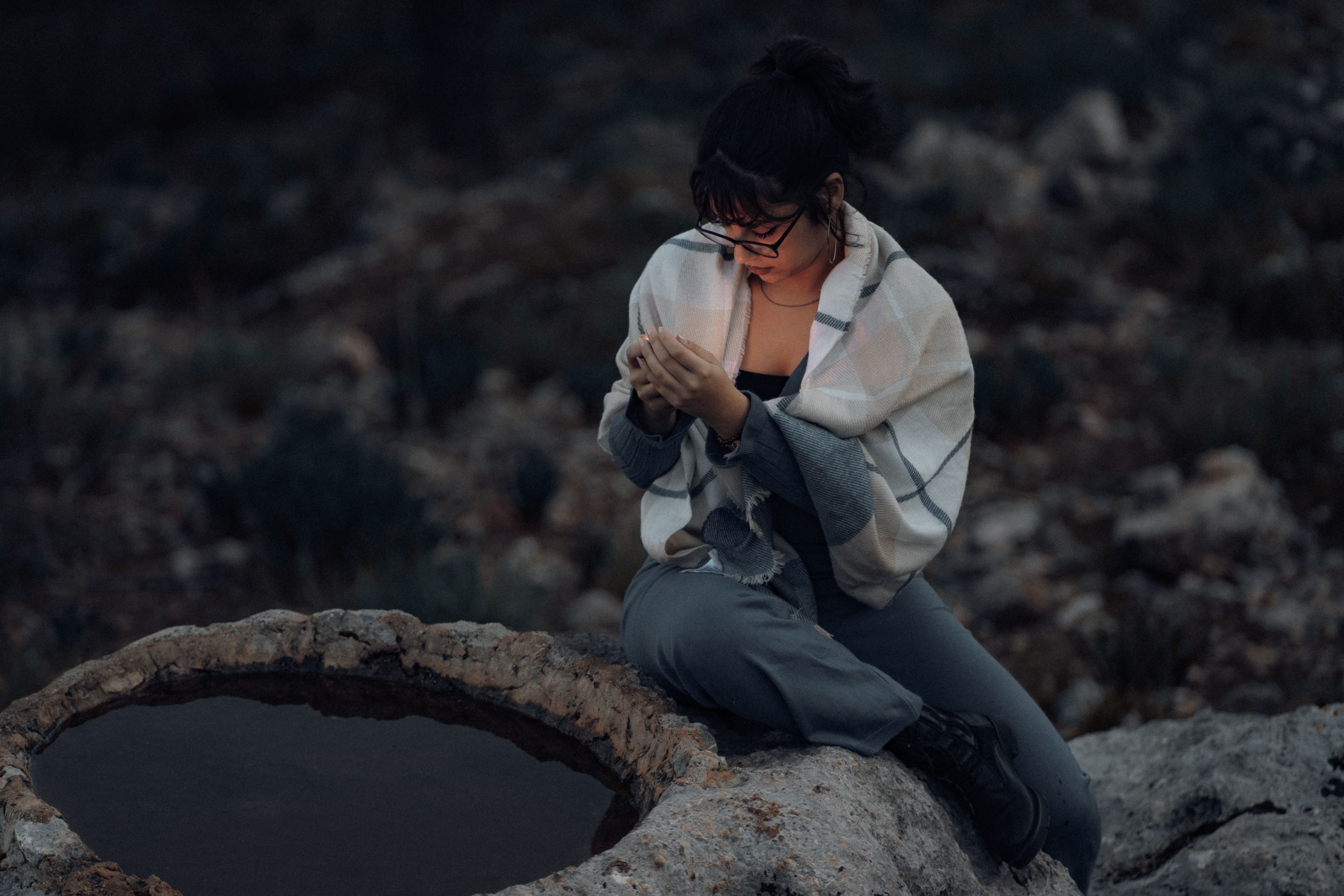 A woman wearing a shawl sits by a rocky pond, looking at her phone.