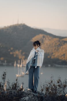 A woman wrapped in a cozy scarf stands by a picturesque lake during sunset.