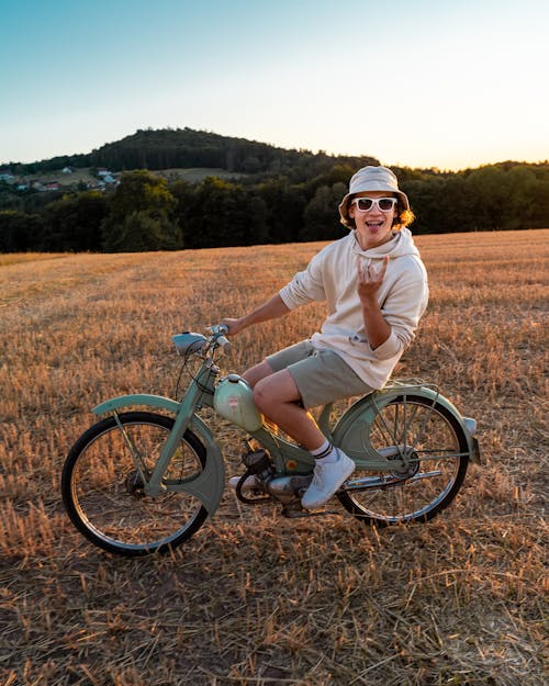 Man in White Hoodie Wearing Sunglasses Riding on a Classic Moped on Brown Grass Field