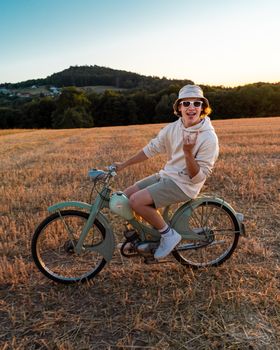 Casual young man enjoying a ride on a vintage moped in a scenic grassy field at sunset.