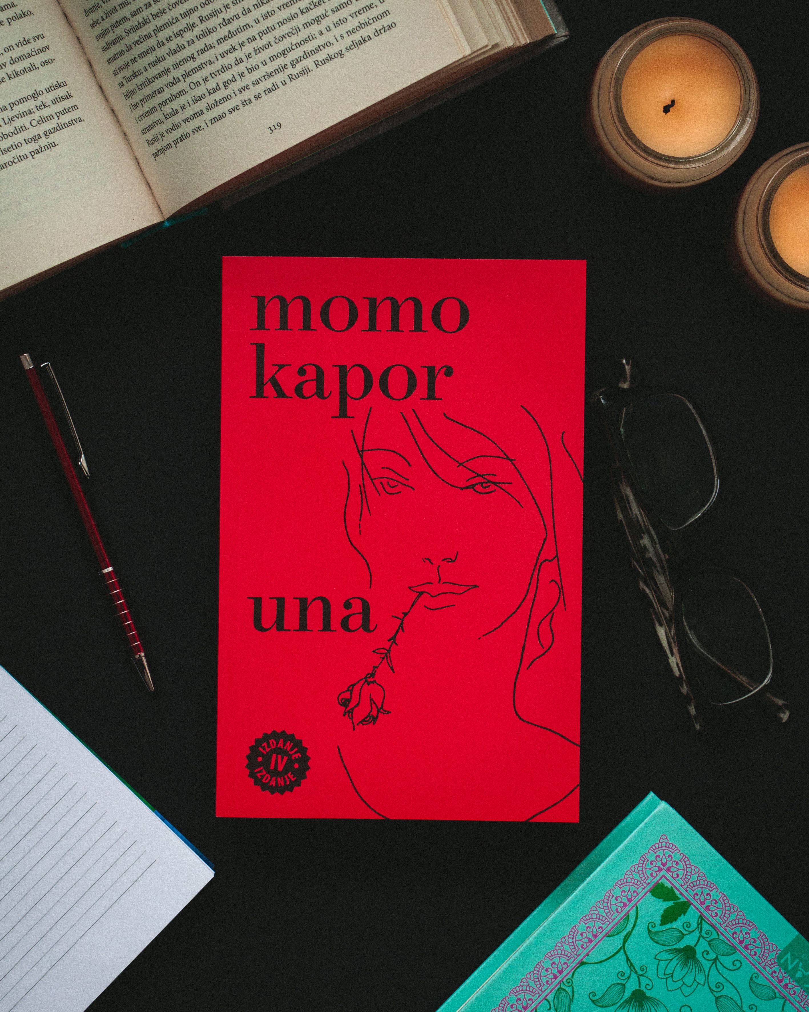 Top view of a red book surrounded by an open notebook, pen, glasses, and candles.