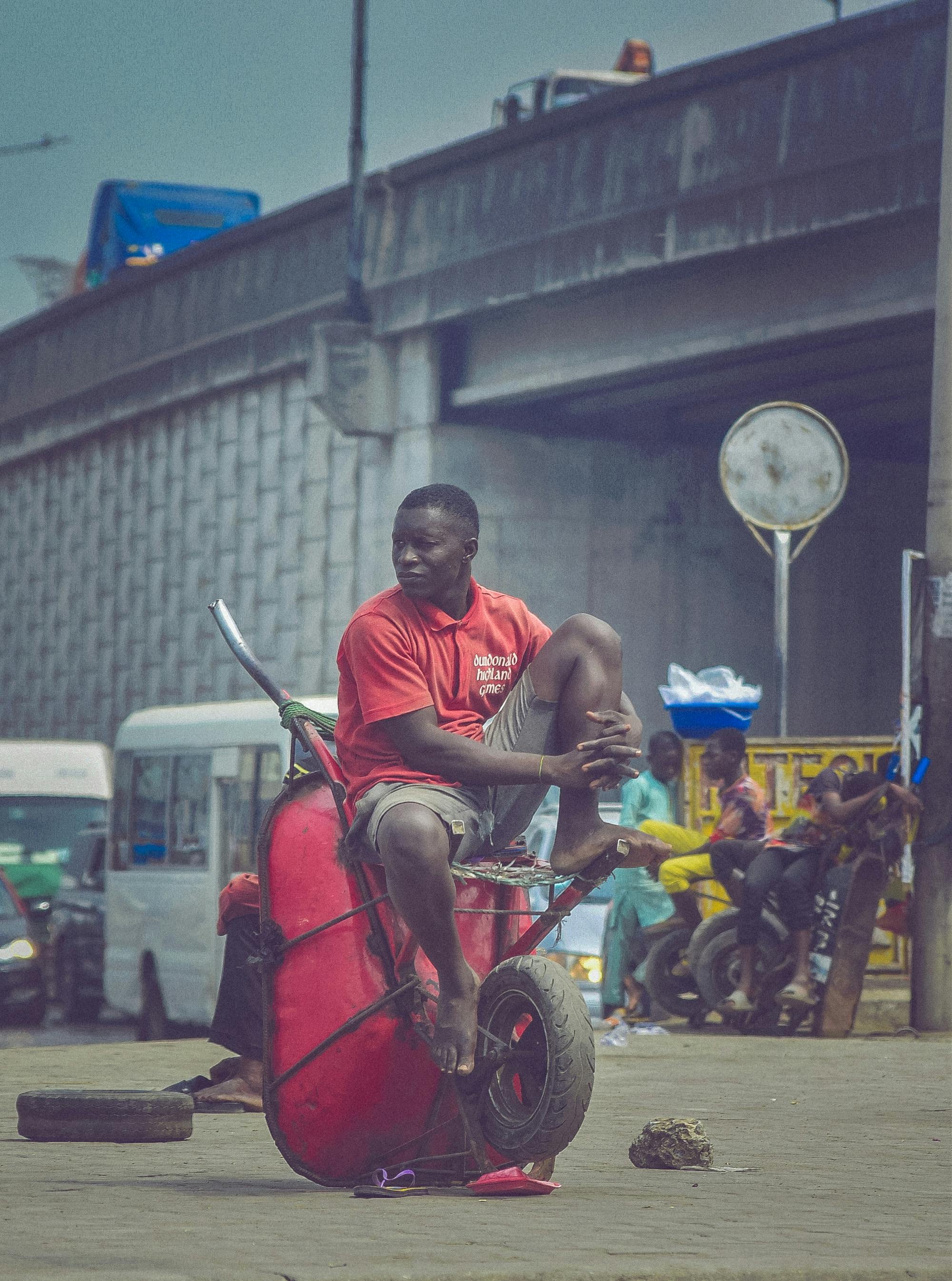 Free Casual urban scene of a man in a red shirt sitting on a wheelbarrow in Lagos, Nigeria. Stock Photo