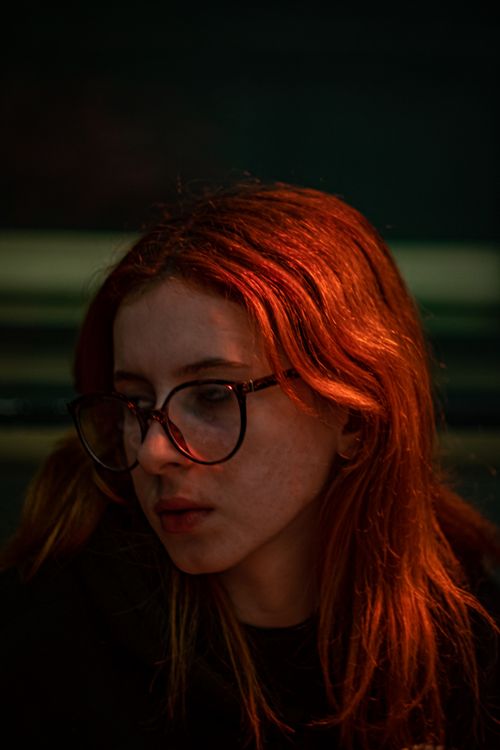 Free A captivating portrait of a woman with vibrant red hair and eyeglasses, captured in a moody ambiance. Stock Photo