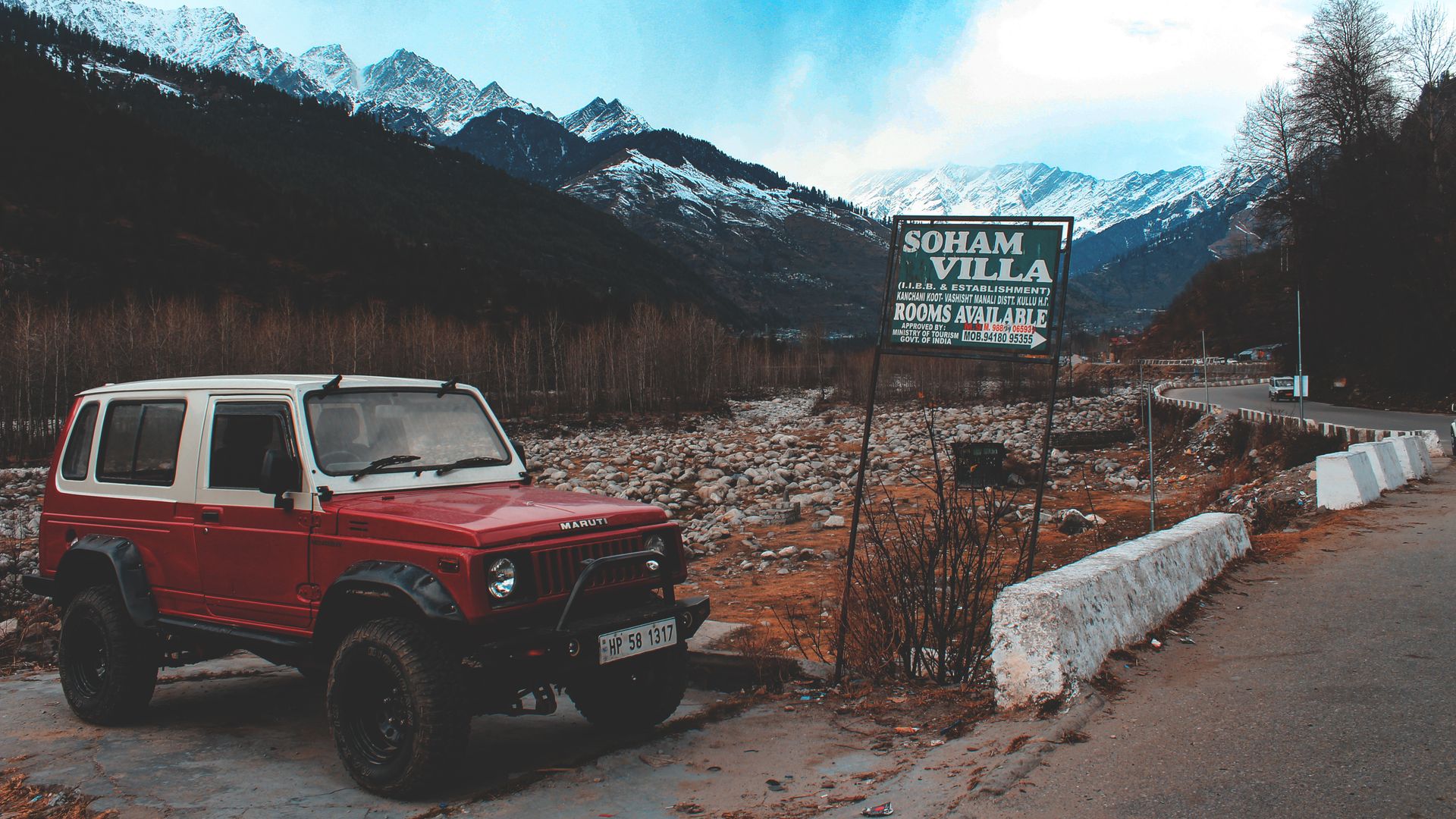 Red SUV parked on a mountain road with snow-capped peaks and a sign for Soham Villa nearby.