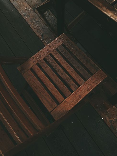 A dark, moody image of a wooden chair covered in rain droplets on a deck.
