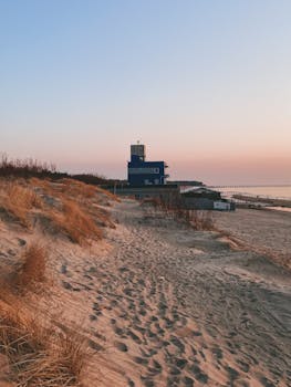 Peaceful coastal scene with sandy dunes, grass, and a blue building at sunset, creating a serene atmosphere.