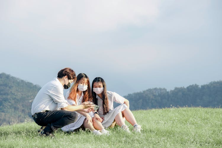 A Group Of Friends Wearing Face Mask Sitting On The Field While Looking At The Phone