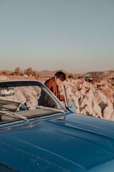 A person stands beside a blue vintage car parked on a scenic cliffside with a vast landscape view.