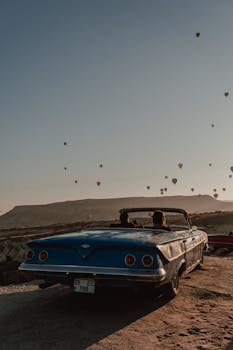 A classic blue convertible with hot air balloons in the sunset sky, creating a nostalgic scene.