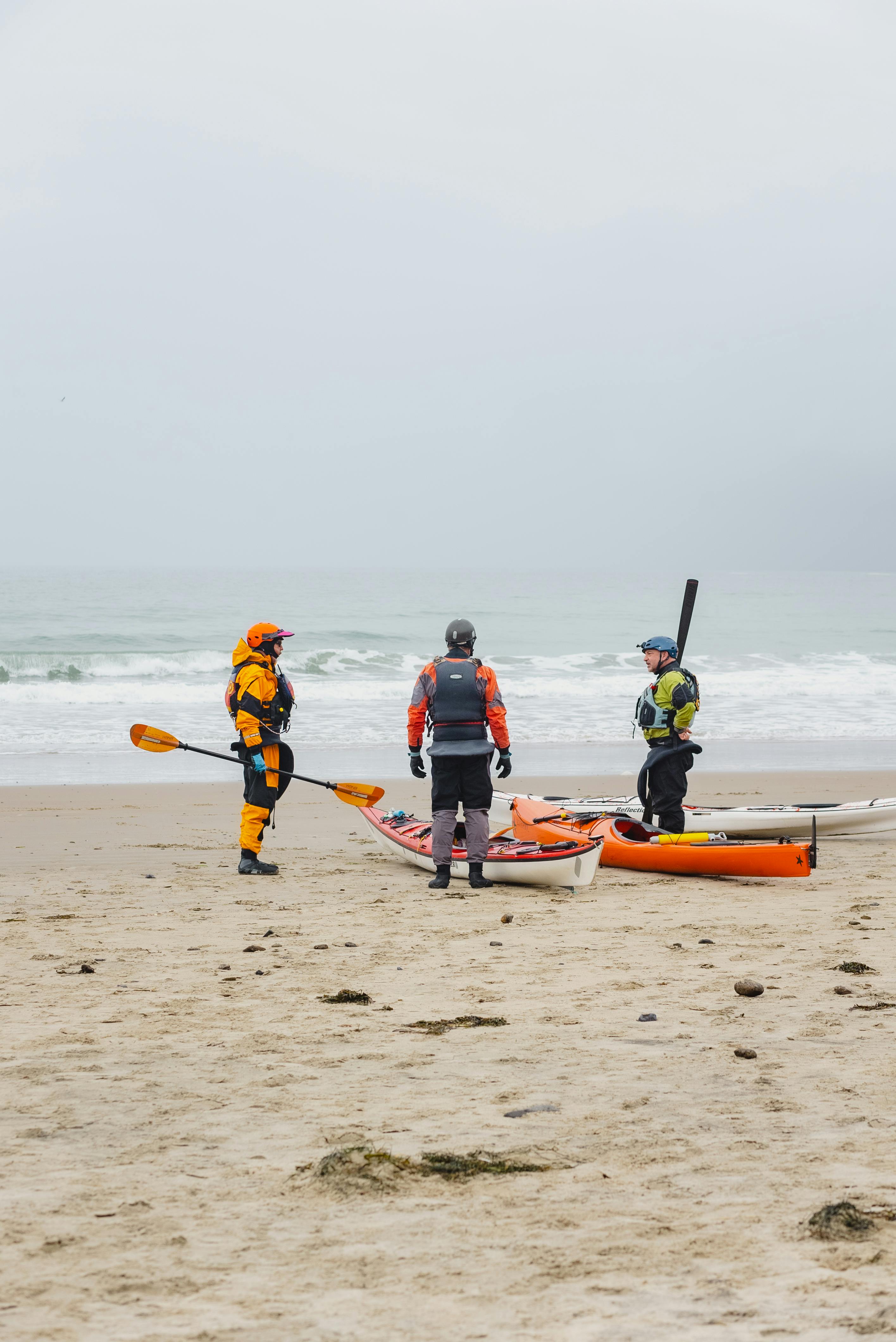 Three adults in gear prepare kayaks on a sandy beach, ready for ocean adventure.
