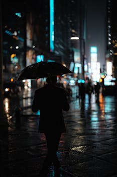 A person with an umbrella walks through a rainy, illuminated urban nightscape, reflecting city lights.