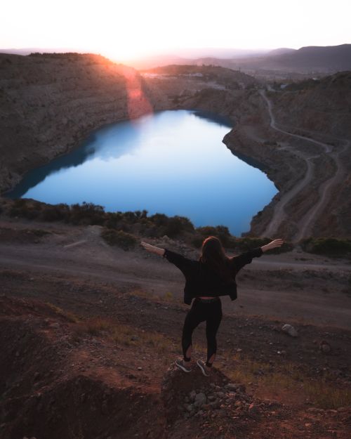 Free Silhouette of a woman standing with open arms overlooking a serene lake during sunset in a rocky landscape. Stock Photo