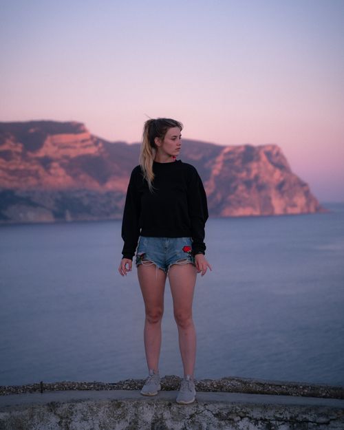Free A young woman in casual attire stands by the sea during sunset, in a picturesque outdoor setting. Stock Photo