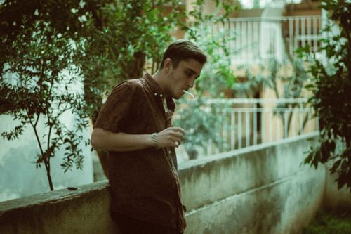 Free A contemplative young man leans against a wall while smoking outdoors. Stock Photo