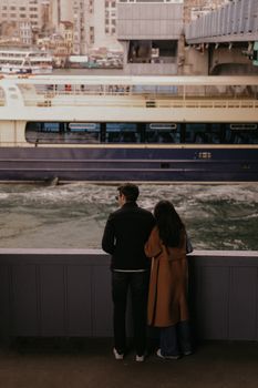 A couple stands by a pier watching a ferry pass by in an urban setting.