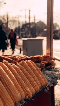 Corn on display at an outdoor market stall with people walking by on a sunny day.