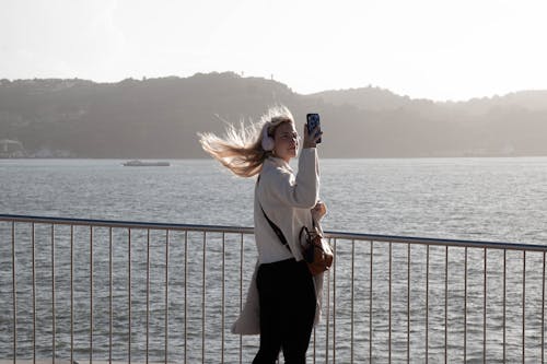 A Woman in White Sweater Standing Near the Ocean while Holding Her Phone