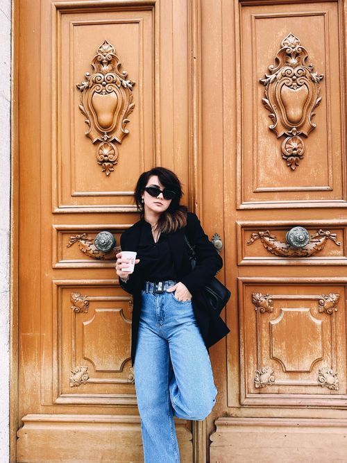 Free Fashion-forward woman posing against a beautifully ornate wooden door with coffee in hand. Stock Photo