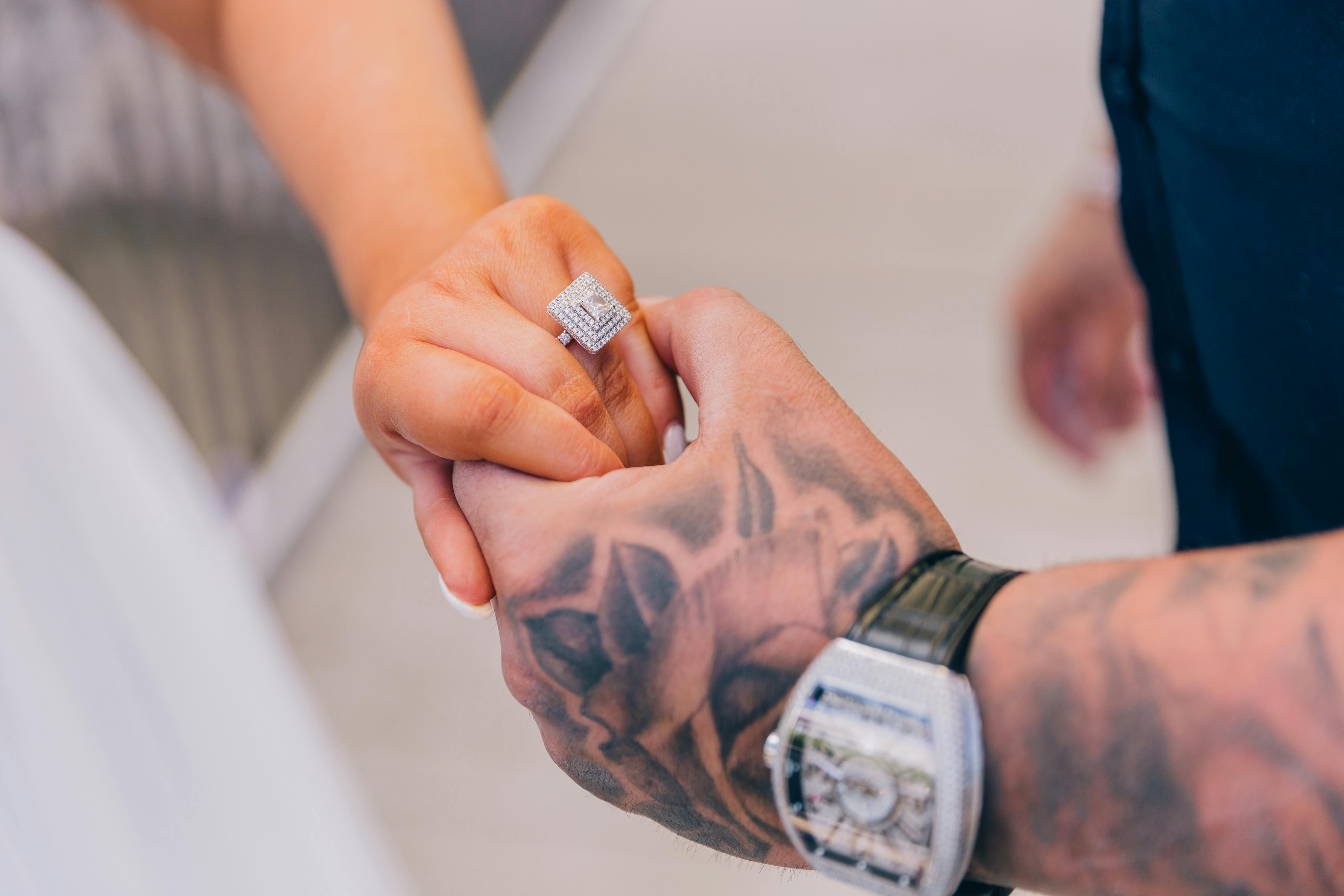 Tender moment of a couple holding hands, featuring a sparkling engagement ring and visible tattoos on the man's arm.