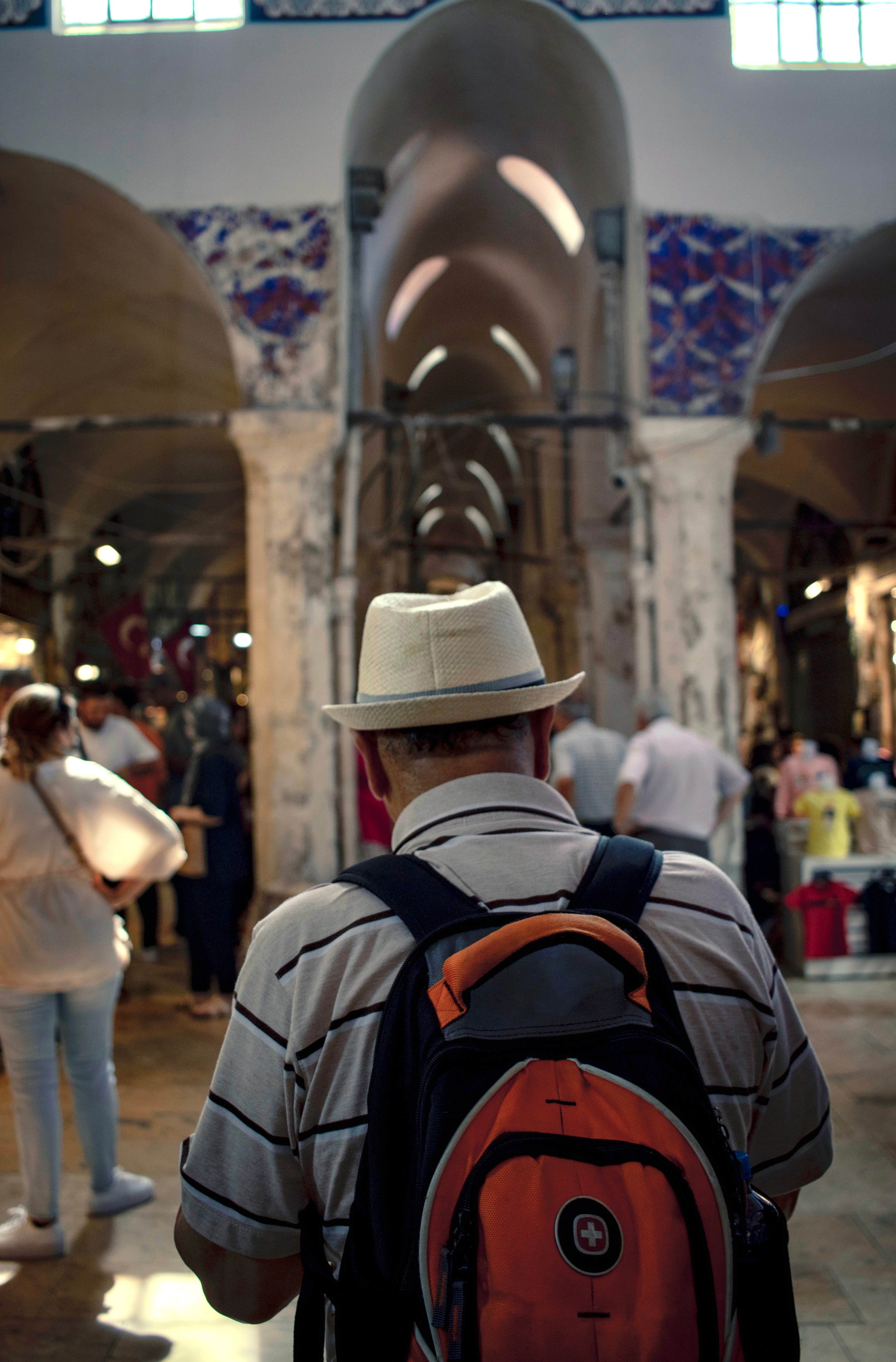 A man with a hat and backpack explores a vibrant indoor market filled with people.