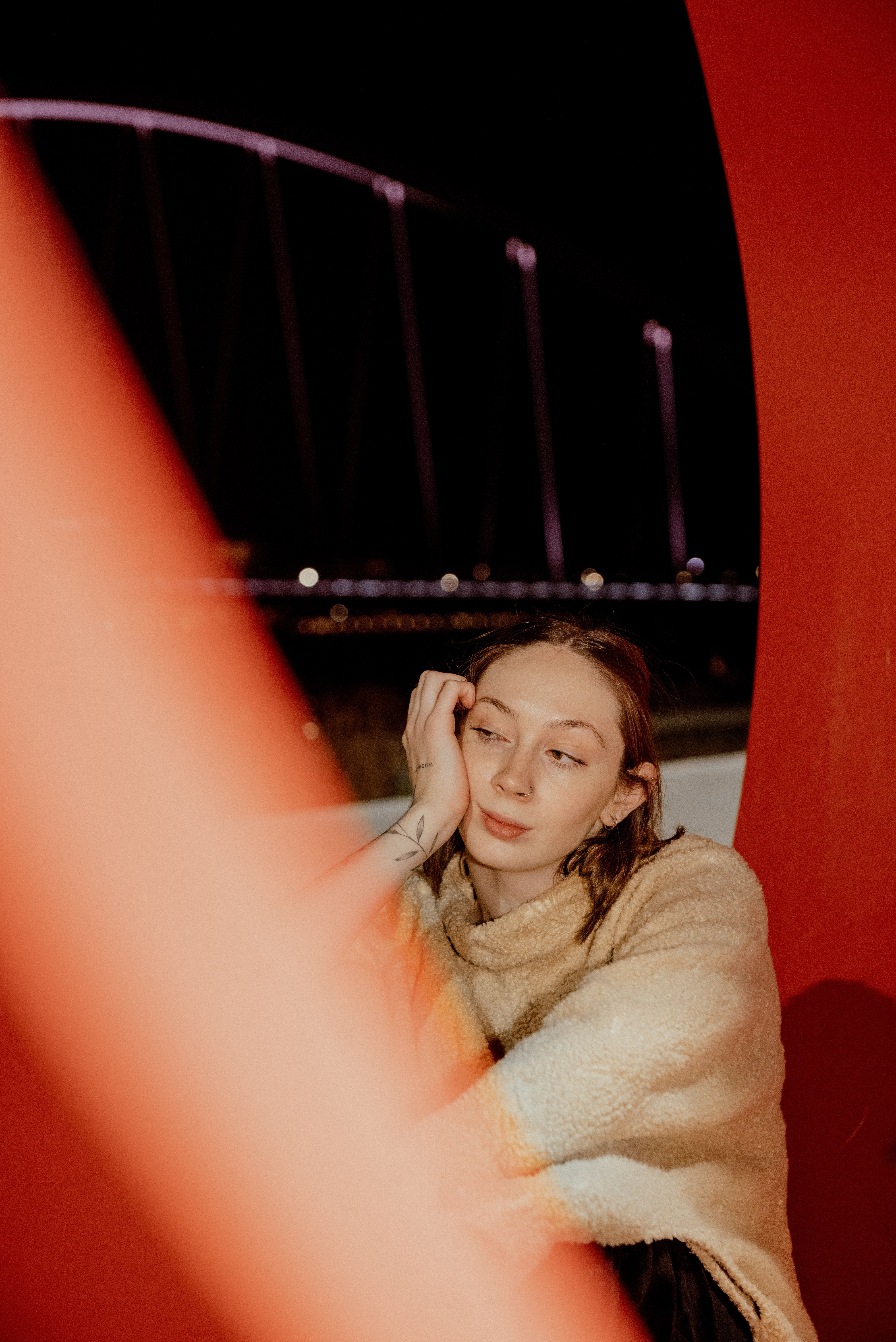 A thoughtful woman in a cozy sweater gazes into the distance, framed by red architecture with an illuminated bridge in the background at night.
