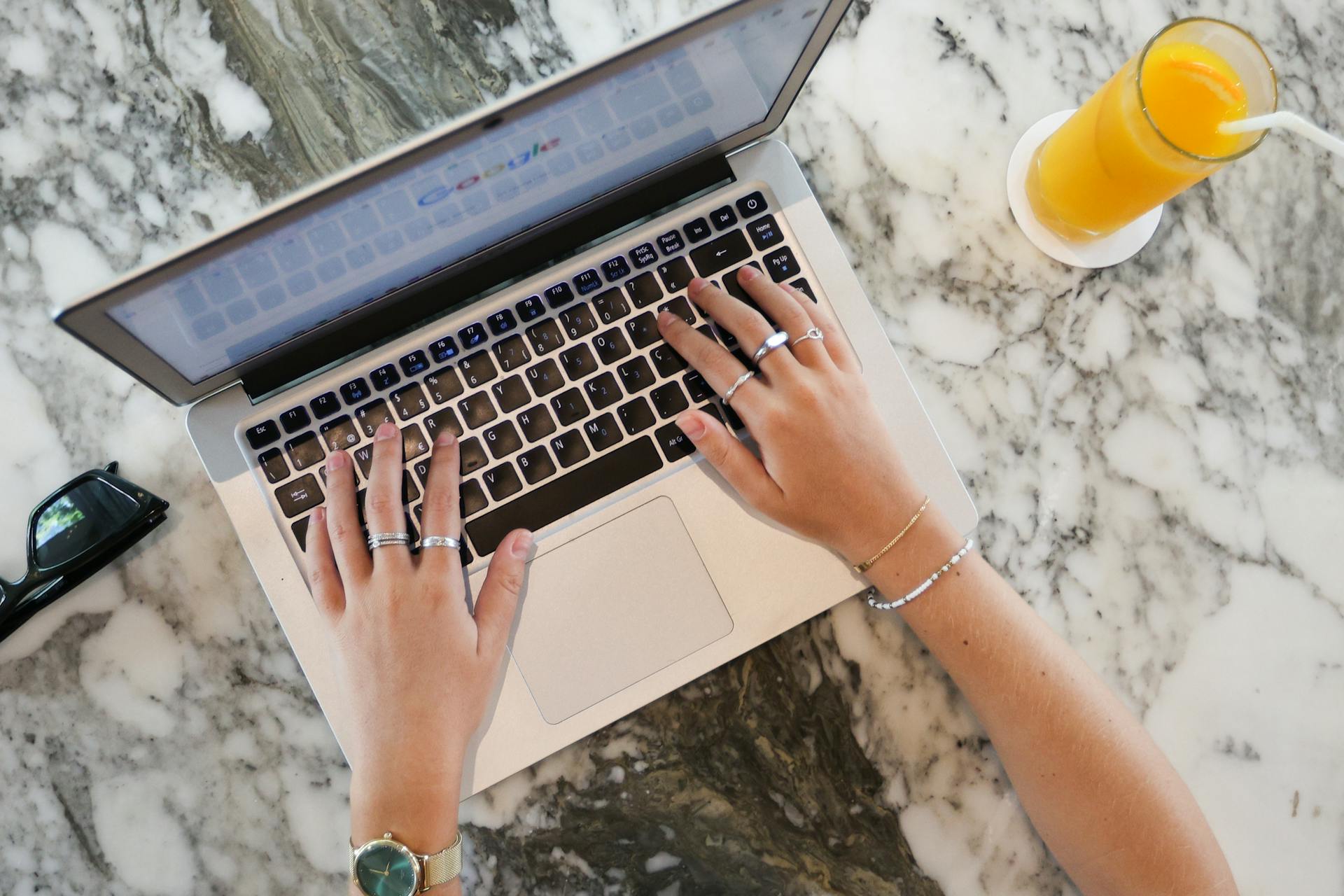 Hands typing on a laptop with orange juice on a marble surface, ideal for remote work themes.